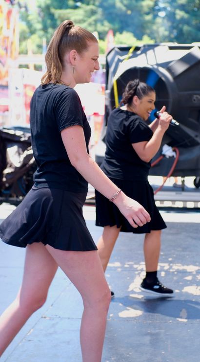 Two performers in black skater skirts dancing and singing on an outdoor festival stage by large speakers, with carnival food booths and crowds in the sunny background.