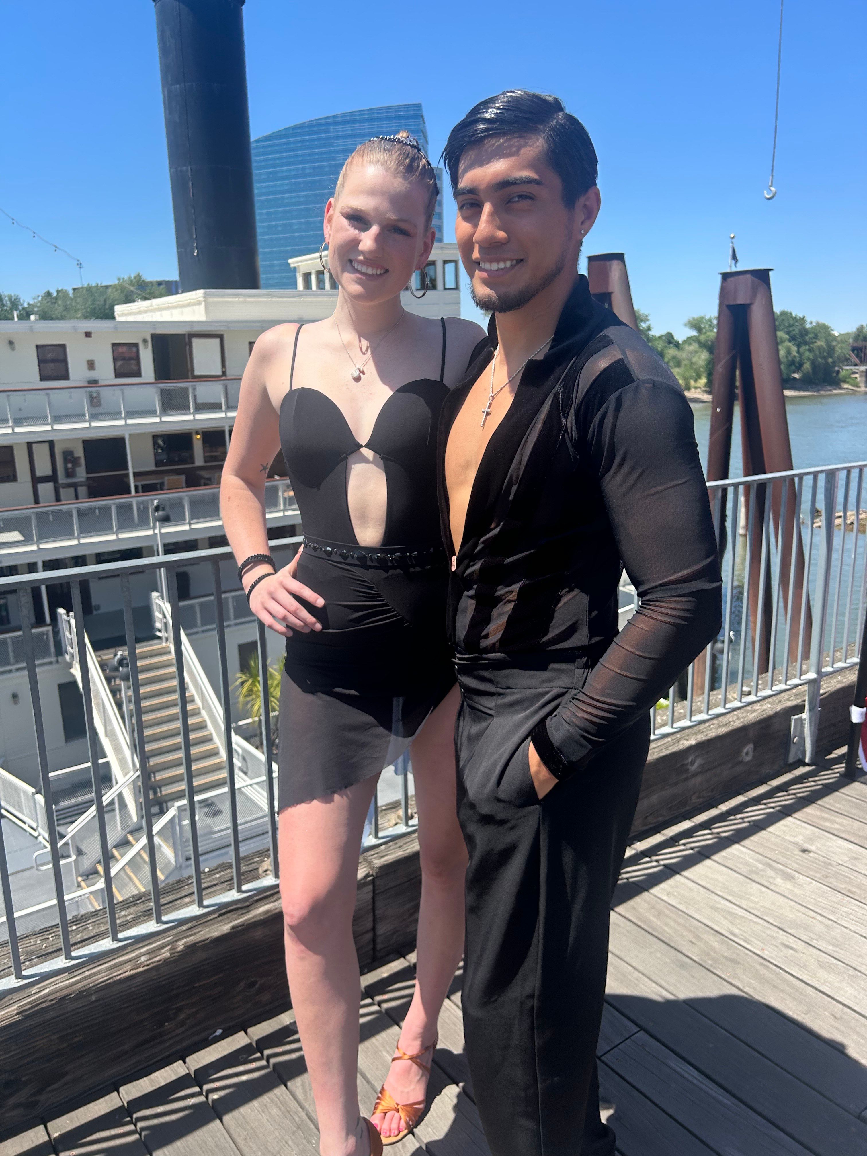 Smiling dance duo in black Latin-style outfits posing on a sunny riverfront deck beside a paddlewheel riverboat and modern glass building in the background.