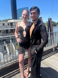 Smiling dance duo in black Latin-style outfits posing on a sunny riverfront deck beside a paddlewheel riverboat and modern glass building in the background.