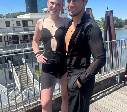 Smiling dance duo in black Latin-style outfits posing on a sunny riverfront deck beside a paddlewheel riverboat and modern glass building in the background.