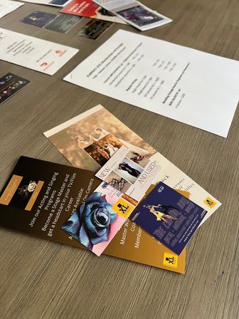 Close-up angled flatlay of colorful brochures, flyers and business cards scattered on a light wood table — wedding promo, event flyer and pricing sheet among tabletop marketing materials.