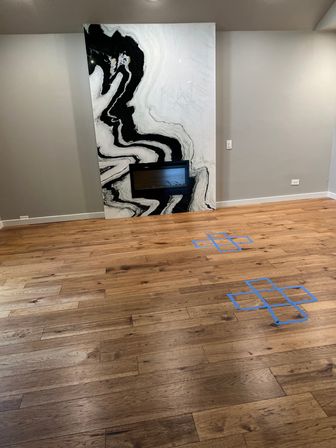 Modern living room interior with warm oak hardwood flooring, large black-and-white marble fireplace panel with built-in electric fireplace, gray walls and blue painter's-tape floor markings
