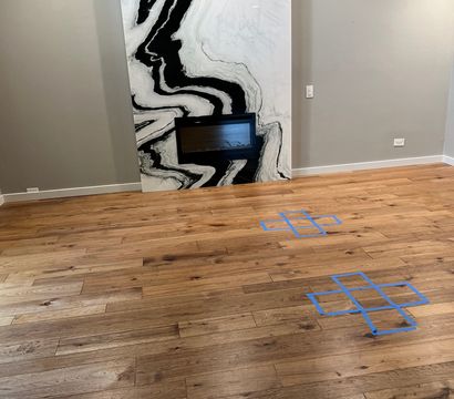 Modern living room interior with warm oak hardwood flooring, large black-and-white marble fireplace panel with built-in electric fireplace, gray walls and blue painter's-tape floor markings