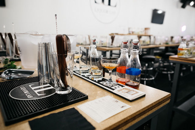 Modern mixology station on a wooden counter with cocktail shakers, bar spoon, tasting flight glasses and small labeled bottles ready for a cocktail tasting class