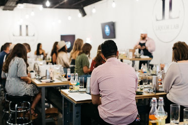 Group cocktail-making class in a bright industrial workshop: participants seated at high tables with bottles, mixers, and glassware while an instructor demonstrates.