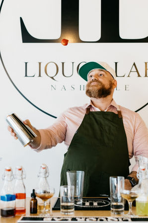 Bearded mixologist in a green apron and cap flipping a small red garnish above a metal shaker at a Nashville cocktail bar counter with bottles and glassware