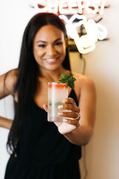 Smiling woman in a black dress holds a frosty cocktail with crushed ice, mint sprig and a splash of red syrup in a tall glass beneath a neon Cheers sign