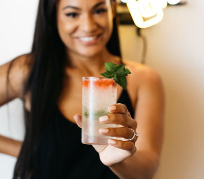 Smiling woman in a black dress holds a frosty cocktail with crushed ice, mint sprig and a splash of red syrup in a tall glass beneath a neon Cheers sign