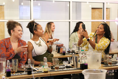 Smiling women at a cocktail bar enjoying a tasting and snapping a selfie over a counter lined with cocktails, bottles and ice buckets