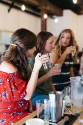 Three people at a cozy bar counter sampling cocktails — one in a red floral off-shoulder dress — holding small tasting glasses amid shakers, an ice bucket and warm string lights overhead.
