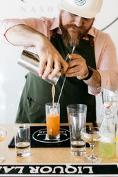 Bartender pouring a bright orange craft cocktail from a metal shaker into a highball glass at a bar counter, surrounded by glassware and mixers — mixology in action.