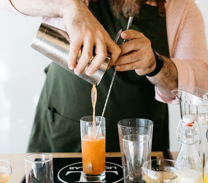 Bartender pouring a bright orange craft cocktail from a metal shaker into a highball glass at a bar counter, surrounded by glassware and mixers — mixology in action.