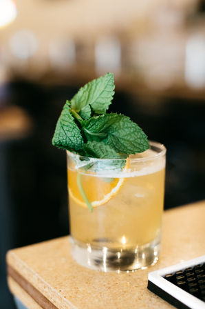 Bright citrus cocktail in a rocks glass with ice, orange peel wheel and a big fresh mint sprig, served on a bar counter