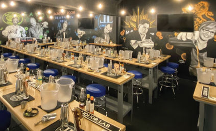 Indoor mixology classroom with rows of wooden workstations set for a cocktail class — shakers, bottles, ice buckets and blue bar stools under hanging bulbs and mural-covered walls.