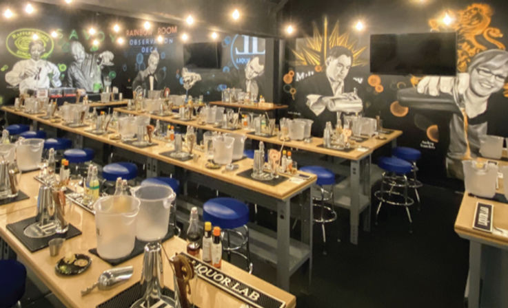 Indoor mixology classroom with rows of wooden workstations set for a cocktail class — shakers, bottles, ice buckets and blue bar stools under hanging bulbs and mural-covered walls.