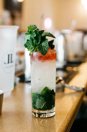 Refreshing tall mint cocktail in a highball glass with crushed ice, muddled mint at the bottom, a red syrup float and a large mint sprig garnish, sitting on a bar counter