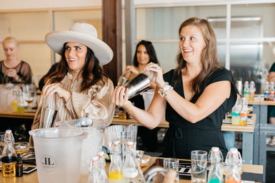 Two smiling women shaking cocktail shakers at a modern mixology class bar counter, surrounded by bottles, glassware and an ice bucket