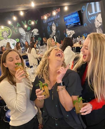 Three friends laughing and holding mint-garnished cocktails at a lively indoor bar with a mural-covered wall, hanging Edison bulbs, and patrons in the background.