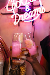 Two hands clinking pink cucumber cocktails beneath a neon "Cocktails & Dreams" sign in a vibrant bar setting