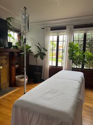Cozy sunlit living room with a covered treatment table, an IV drip on a pole, hardwood floors and potted plants by tall windows.