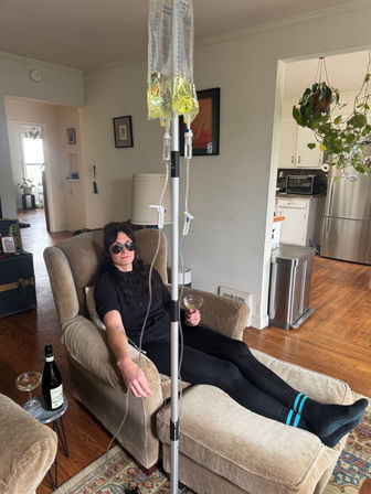 Person lounging in a cozy living-room armchair wearing sunglasses, connected to a home IV drip with two yellow bags and sipping a cocktail.