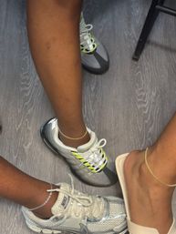 Three feet on gray wood floor — two in sporty sneakers (metallic silver and gray with neon yellow accents) and one in a beige slide, each wearing delicate anklets.