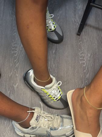 Three feet on gray wood floor — two in sporty sneakers (metallic silver and gray with neon yellow accents) and one in a beige slide, each wearing delicate anklets.
