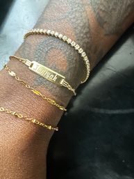 Close-up of a tattooed wrist wearing a gold nameplate bracelet, a sparkling tennis-style bracelet and two delicate gold chains against a dark background.