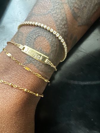 Close-up of a tattooed wrist wearing a gold nameplate bracelet, a sparkling tennis-style bracelet and two delicate gold chains against a dark background.