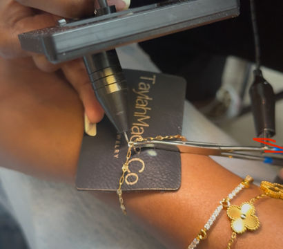 Close-up of a jeweler's hand using a handheld tool and pliers to fasten a delicate gold chain to a bracelet on a black jewelry tag, wrist wearing layered gold charm bracelets.