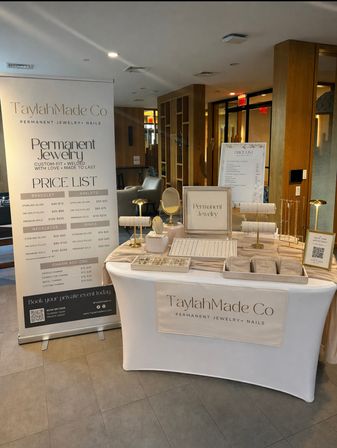 Pop-up jewelry kiosk in a modern lobby with a white-draped table, gold-accent display stands, trays of delicate bracelets and necklaces, and a tall price-list banner advertising permanent jewelry.
