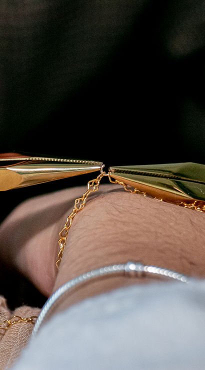 Close-up of hands with pink lace nail art using gold pliers to fasten a delicate gold chain bracelet clasp on a wrist.
