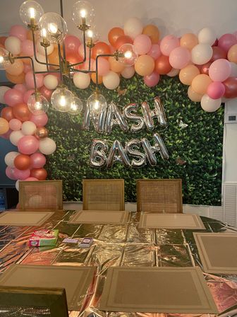 Indoor party decor with pink, peach and rose-gold balloon garland on a faux green leaf wall, silver foil letter balloons reading "MASH BASH", a modern glass-globe chandelier, and a dining table set with placemats.
