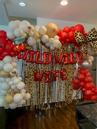 Indoor party backdrop with red foil letter balloons reading WILD WILD WIFE, red and white balloon arch, leopard-print bow and gold fringe curtain for a playful celebration.