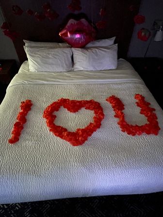 Romantic hotel bed with white quilt, red rose petals arranged to read 'I ♥ U', red lips balloon and hanging flower decorations