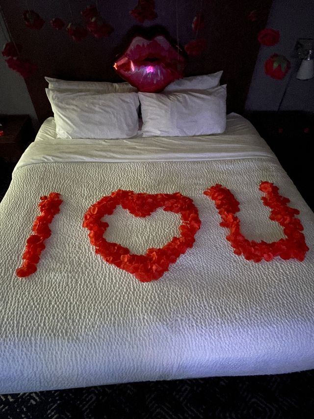 Romantic hotel bed with white quilt, red rose petals arranged to read 'I ♥ U', red lips balloon and hanging flower decorations