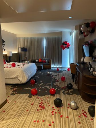 Romantic hotel suite decorated with red, black and silver balloons and scattered red rose petals across the bed and carpet, cozy sofa and desk by a curtained window.