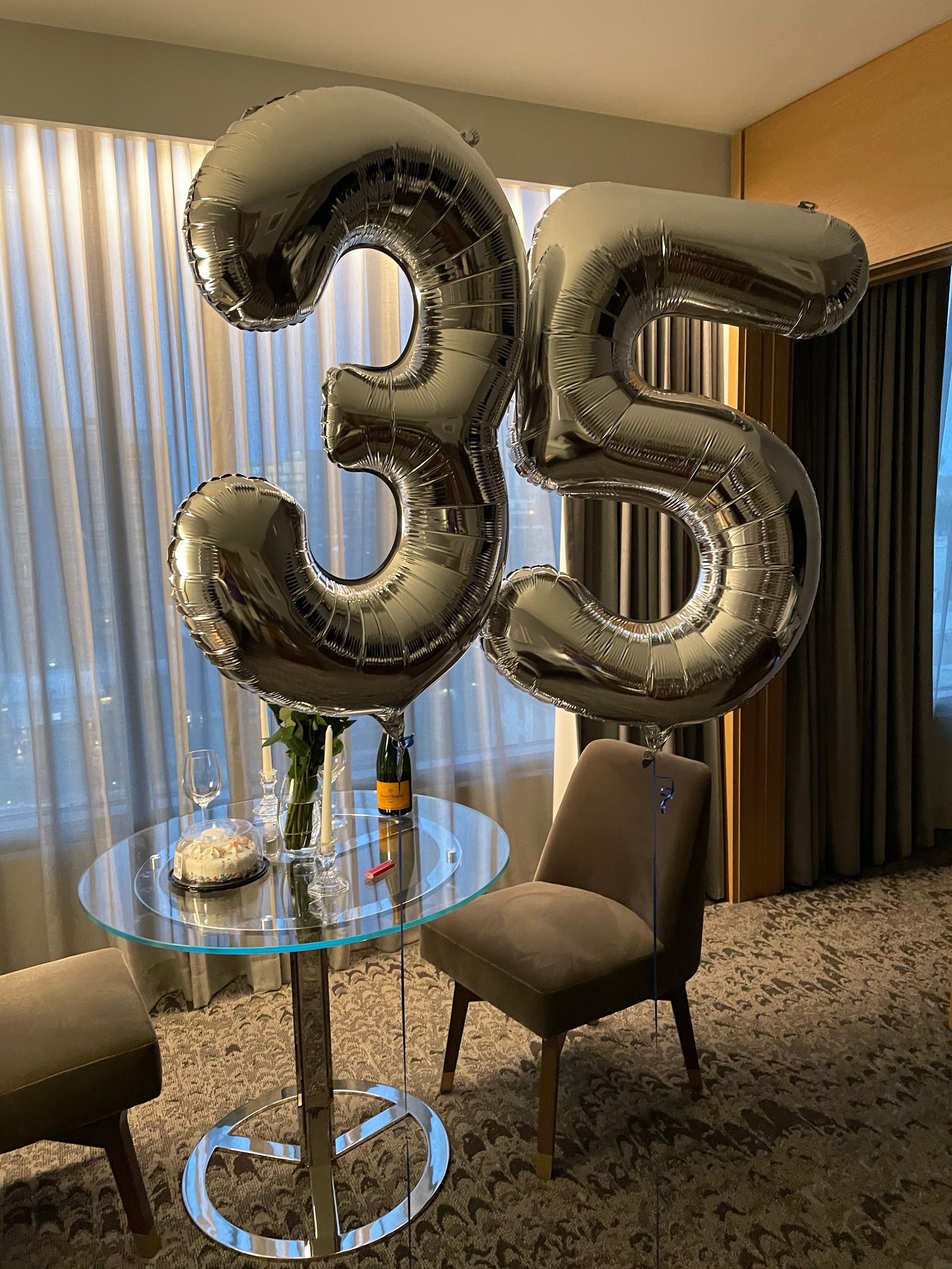 Silver foil '35' balloons floating above a glass table in a hotel suite, with a champagne bottle, small cake, two chairs and floor-to-ceiling curtains framing a city view.