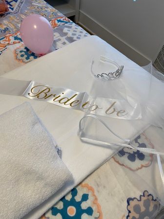 Bride-to-be satin sash with gold script, rhinestone tiara and veil laid on a patterned bed with a pink balloon — bachelorette/bridal shower prep