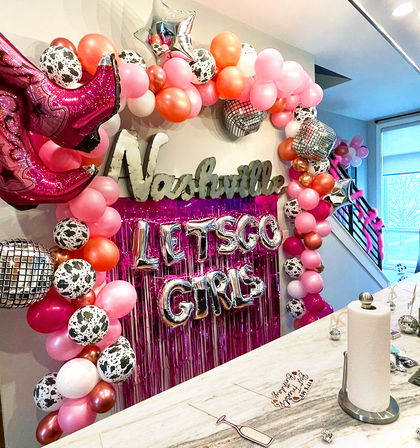 Nashville bachelorette party balloon backdrop with pink and rose-gold balloon arch, cow-print and disco ball balloons, foil cowboy boot and silver "LET'S GO GIRLS" letters over pink fringe above a kitchen island.