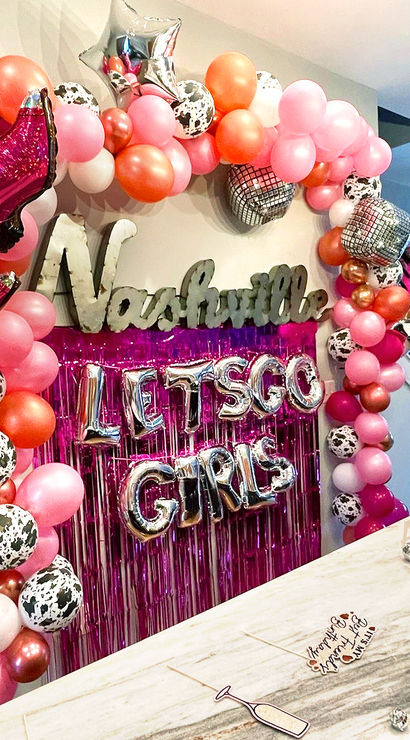 Nashville bachelorette party balloon backdrop with pink and rose-gold balloon arch, cow-print and disco ball balloons, foil cowboy boot and silver "LET'S GO GIRLS" letters over pink fringe above a kitchen island.