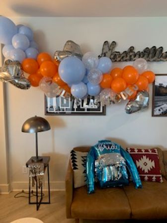 Indoor living room party scene with a blue, orange and silver balloon garland above framed photos, a shiny teal sequin party jacket draped over a tan sofa with patterned pillows and a modern floor lamp nearby.