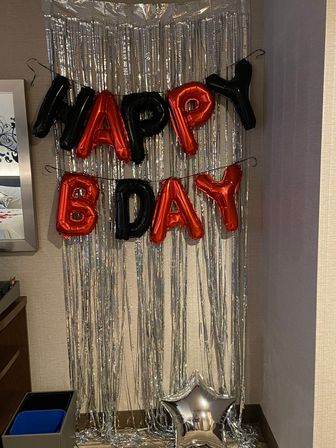 Red and black foil 'HAPPY BDAY' balloons on a silver fringe curtain party backdrop with a reflective silver star balloon on the floor in a hotel room corner