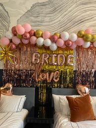 Bridal shower bedroom backdrop with pink, white and gold balloon garland, rose-gold foil fringe and metallic 'BRIDE' and 'love' balloons above two beds with heart-shaped foil accents