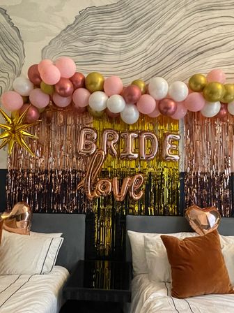 Bridal shower bedroom backdrop with pink, white and gold balloon garland, rose-gold foil fringe and metallic 'BRIDE' and 'love' balloons above two beds with heart-shaped foil accents