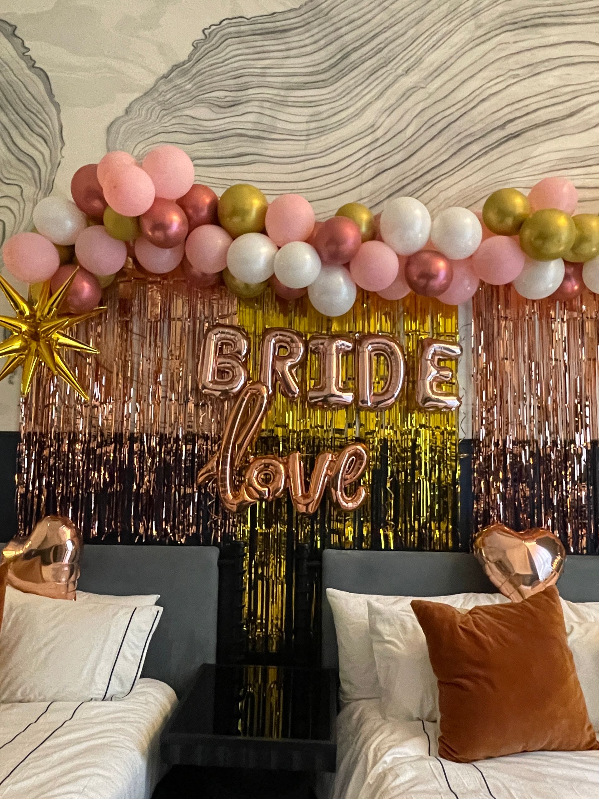 Bridal shower bedroom backdrop with pink, white and gold balloon garland, rose-gold foil fringe and metallic 'BRIDE' and 'love' balloons above two beds with heart-shaped foil accents