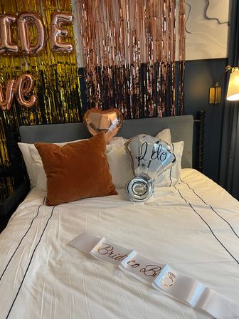 Bridal suite bed styled for a bachelorette with rose-gold fringe backdrop, metallic heart and “I do” balloons, rust velvet pillow and a “Bride to Be” sash on white linens.