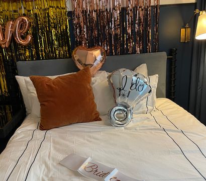 Bridal suite bed styled for a bachelorette with rose-gold fringe backdrop, metallic heart and “I do” balloons, rust velvet pillow and a “Bride to Be” sash on white linens.