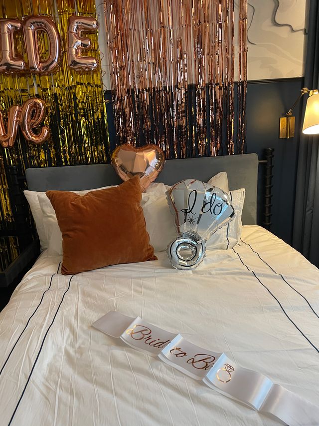 Bridal suite bed styled for a bachelorette with rose-gold fringe backdrop, metallic heart and “I do” balloons, rust velvet pillow and a “Bride to Be” sash on white linens.
