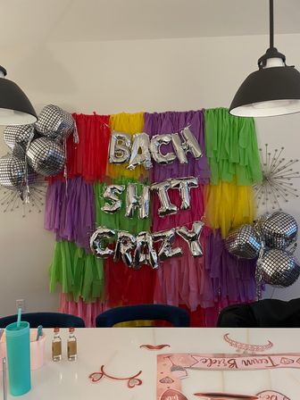 Indoor bachelorette party scene with rainbow tissue tassel backdrop, silver foil letter balloons spelling a playful phrase, mirrored disco-ball balloons, hanging pendant lights and a table of party props and drinks.